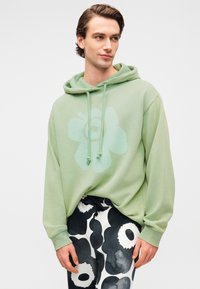 Φούτερ σε ανοιχτό πράσινο με μεγάλο σχέδιο λουλουδιού στο ίδιο χρώμα. Διαθέτει ρυθμιζόμενα κορδόνια και μακριά μανίκια, σε συνδυασμό με παντελόνια μαύρου και άσπρου με σχέδιο.