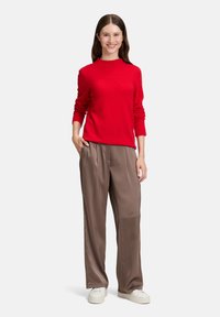 Kobieta stoi i się uśmiecha, ubrana w czerwoną bluzę, luźne brązowe spodnie i białe trampki, z jedną ręką w kieszeni.