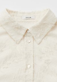 Blusa crema con collo, superficie strutturata, dettagli sfrangiati, chiusura con bottoni e etichetta del marchio OPUS sul collo.