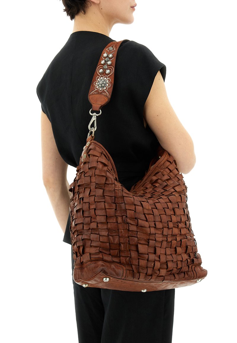 Donna con top nero senza maniche che porta una grande borsa a tracolla in pelle intrecciata marrone con tracolla decorata a rilievo floreale e borchie metalliche alla base.