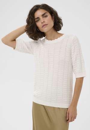 Jeune femme aux cheveux bruns courts et bouclés, portant un pull blanc texturé à manches courtes et une jupe beige, debout devant un fond uni.