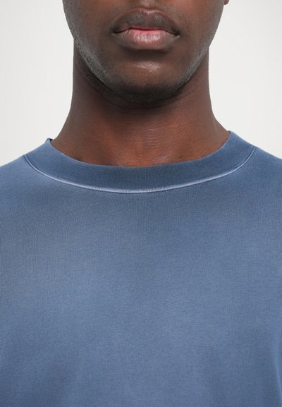 Modra majica s krojem okroglega vratu, izdelana iz gladkega, lahkega materiala. Ima rahlo rebrasto teksturo, brez vidnih vzorcev ali poudarkov.