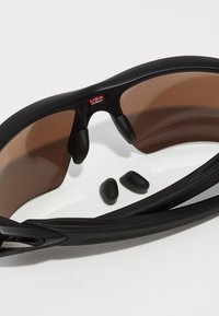 Gafas de sol deportivas negras con lentes marrones, almohadillas de nariz de goma y agarraderas laterales. Incluye dos piezas de acento de goma negra adicionales.