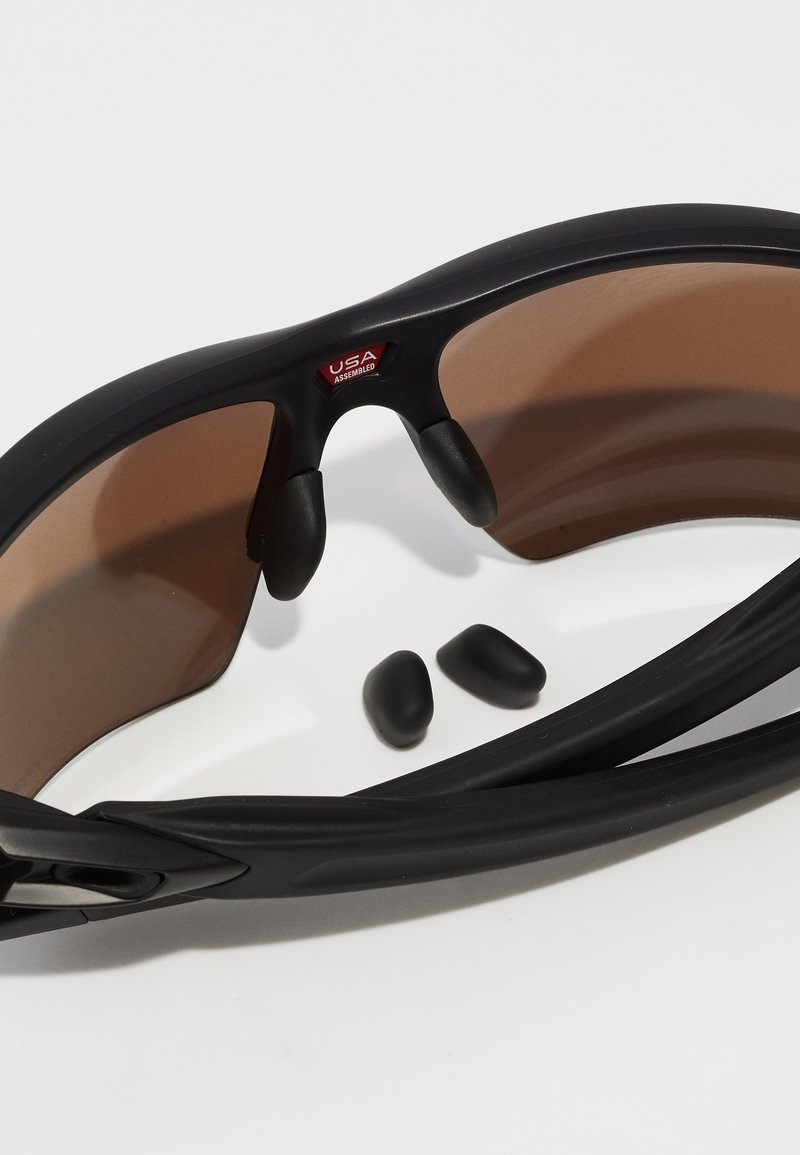 Schwarze Sportsonnenbrille mit braunen Gläsern, Gummiauflagen an der Nase und seitlichen Griffen. Enthält zwei zusätzliche schwarze Gummibezogene Teile.