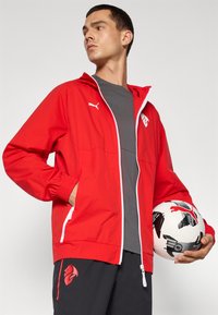 Rød letvægtsjakke med hætte, som har en frontlynlås og elastiske manchetter, parret med sorte shorts og en mønstret fodbold.