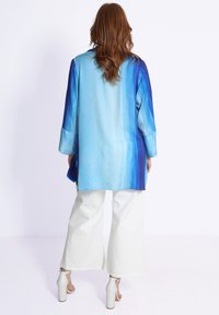 Chemise légère à manches longues dégradée bleue avec une coupe ample, accompagnée d'un pantalon large blanc et de sandales. Texture lisse, design décontracté.