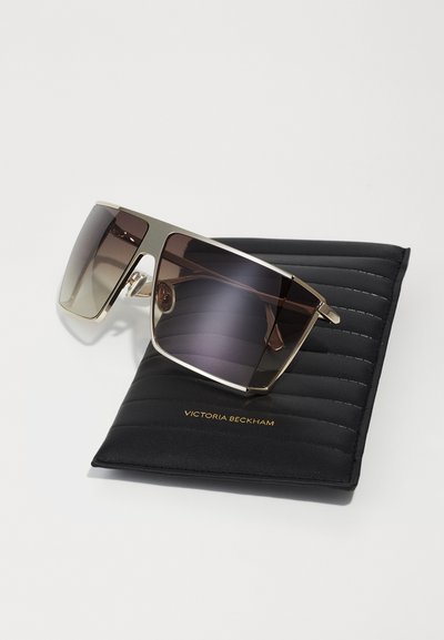 Lunettes de soleil avec une monture rectangulaire et des verres dégradés marron, des accents dorés, reposant sur un étui noir matelassé avec un texte en relief.