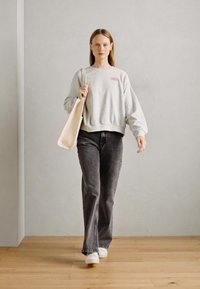 Grijze sweatshirt met geribbelde manchetten, grafisch detail op de borst, gecombineerd met donkergrijze wijde jeans en witte sneakers. Beige schoudertas in de hand.