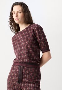 Strickoberteil mit kurzen Ärmeln, das ein weinrotes Diamantmuster mit goldenen Akzenten aufweist. Gerippter Halsausschnitt und Saum, strukturiert mit einem subtilen Schimmer.