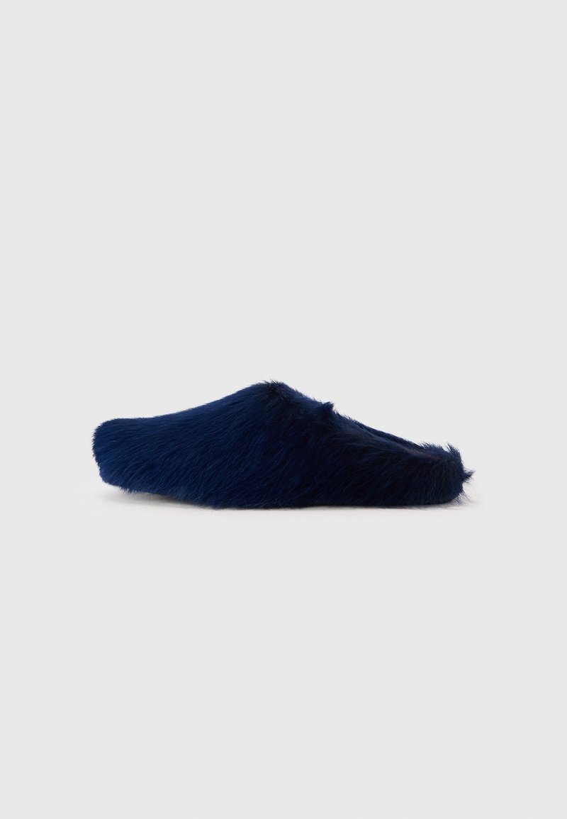 Chausson en fausse fourrure bleu marine à enfiler, avec un bout fermé et un dos ouvert, semelle plate, isolé sur un fond clair.
