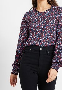 Femme portant un chemisier à manches longues à fleurs avec des poignets à volants et un pantalon noir taille haute, sur un fond blanc uni.