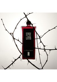 Bottiglia di profumo in vetro rosso con un'etichetta nera su cui è inciso "Serge Lutens". Circondata da spine scure e acuminate su uno sfondo chiaro.