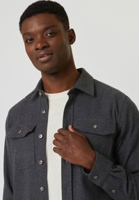 Giacca in lana grigia con bottoni, due tasche sul petto e bottoni a contrasto, indossata sopra una camicia chiara a macchie.