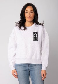 Sweatshirt gris clair avec un col rond, coupe oversize et manches longues. Présente un design graphique noir sur le devant.