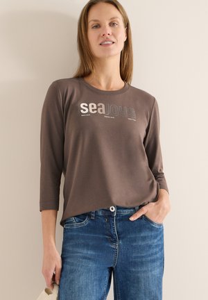 Brun långärmad T-shirt med texten "sea JOYS" i ljusa färger, kombinerad med blå jeans. Mjukt tyg med avslappnad passform.