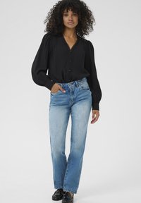 Vrouw met krullend haar, gekleed in een zwarte blouse met lange mouwen, lichtblauwe spijkerbroek en zwarte schoenen, staand met één hand in haar zak tegen een witte achtergrond.