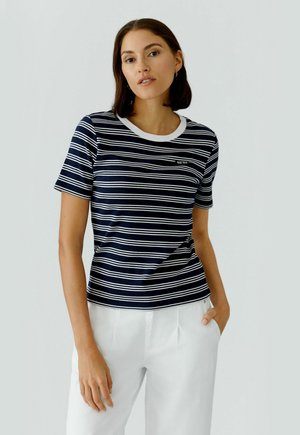 Frau mit kurzen braunen Haaren, die ein marineblau-weiß gestreiftes kurzärmeliges Hemd und weiße Hosen trägt, steht mit einer Hand in der Tasche.