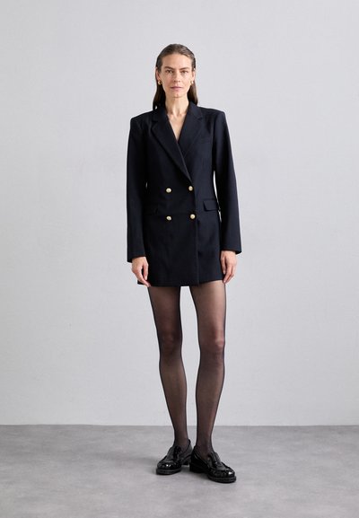 Marineblå dobbeltspent blazer med gullknapper, notched lapels, to frontlommer; kombinert med svarte strømpebukser og loafers.
