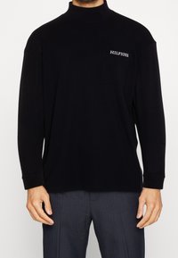 Mies, kuris dėvi juodą ilgą rankovę marškinius su krūtinės kišene, pažymėta "HILFIGER", ir tamsiai dryžuotas kelnes.