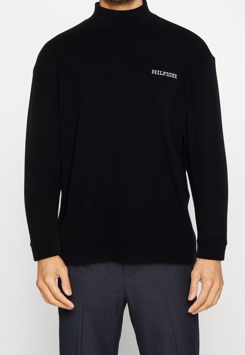 Mies, kuris dėvi juodą ilgą rankovę marškinius su krūtinės kišene, pažymėta "HILFIGER", ir tamsiai dryžuotas kelnes.