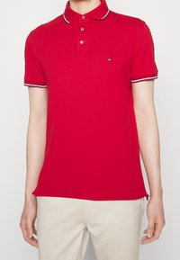 Homme portant un polo rouge ajusté avec un col boutonné et une bordure rayée sur les manches et le col, associé à un pantalon beige.