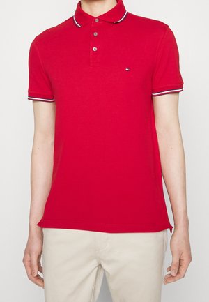 Homme portant un polo rouge ajusté avec un col boutonné et une bordure rayée sur les manches et le col, associé à un pantalon beige.