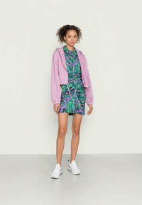 Moletom cropped de lavanda sobre um vestido estampado em verde e roxo com detalhe de cordão. Modelo usa tênis brancos, de pé sobre um fundo neutro.