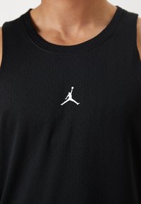 Débardeur de sport noir avec une texture en mesh respirant, présentant un logo Jordan blanc au niveau de la poitrine. Tissu lisse avec un col rond.
