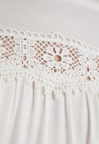 Vit bomullsskjorta med detaljerad spetskant med blommönster och spiralformer, strukturerad med en mjuk yta och subtila veck under spetsen.