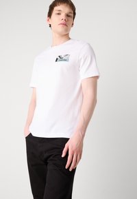 Balta kokvilnas T-kreklu ar grafisku kabatas detaļu un "Calvin Klein" etiņu. Īsas piedurknes, apaļa izgriezuma kakla daļa, gluda tekstūra.