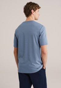 Jonge man van achteren gezien, gekleed in een lichtblauw shirt met korte mouwen en een donkere broek, met handen in de zakken tegen een effen achtergrond.