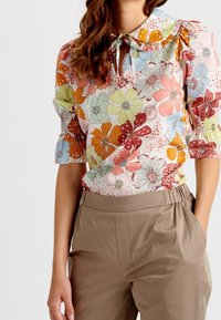 Blusa floreale in colori pastello con maniche a sbuffo, colletto arricciato e apertura a occhiello. Indossata con pantaloni beige a vita alta.
