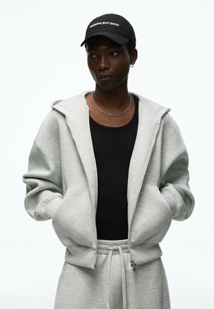 Junge Person trägt eine schwarze Kappe mit "Nothing But Good," grauem Kapuzenpullover mit Reißverschluss, schwarzem Tanktop, silberner Kette und grauer Jogginghose, Hände in den Taschen.