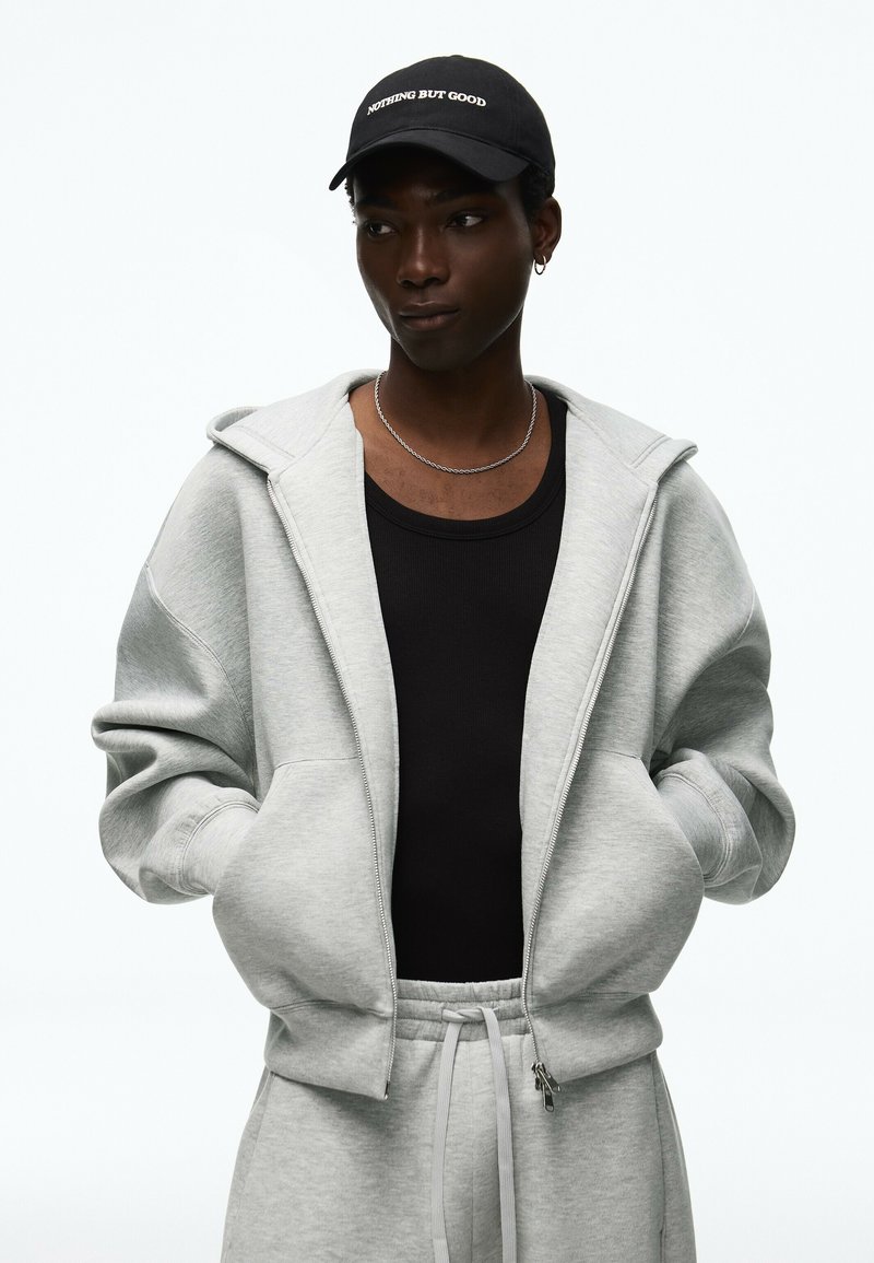 Jeune personne portant une casquette noire avec un sweat à capuche gris zippé "Nothing But Good,", un débardeur noir, une chaîne argentée et un pantalon de survêtement gris, mains dans les poches.