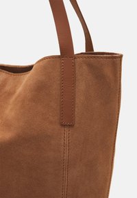Sac fourre-tout en daim marron avec des accents en cuir lisse, double poignées et coutures visibles le long des joints. Texture douce avec un design simple.