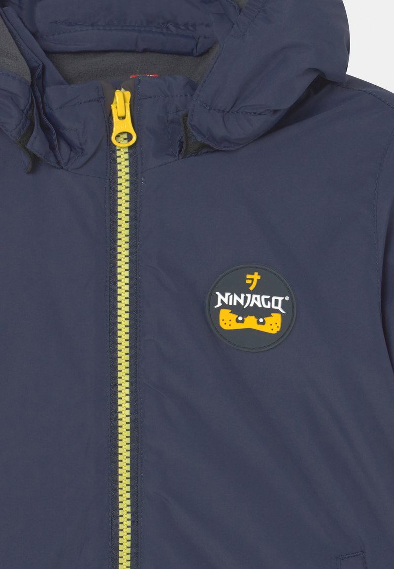 Marineblaue Jacke mit Kapuze, gelbem Reißverschluss und einem runden schwarzen Aufnäher mit dem Ninjago-Logo in Gelb und Weiß. Glatte Textur.