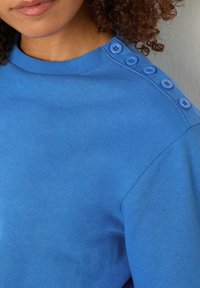 Felpa blu con un collo a coste e sei bottoni blu decorativi sulla spalla sinistra, caratterizzata da una superficie liscia e uniforme.