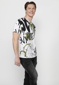 Camiseta blanca de manga corta con patrones de salpicaduras en negro y verde oliva, y texto en estilo grafiti, combinada con pantalones cargo oscuros y texturizados.