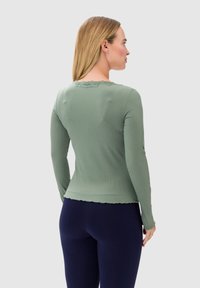 Maglia a maniche lunghe, a coste, di morbido verde oliva con scollo in pizzo a scallop. Abbinata a leggings scuri, che evidenziano un silhouette aderente.
