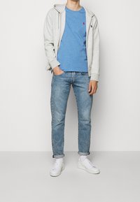 Gray zip-up hoodie over a blue t-shirt, paired with light blue jeans and white sneakers. Simple, casual outfit with a relaxed fit.