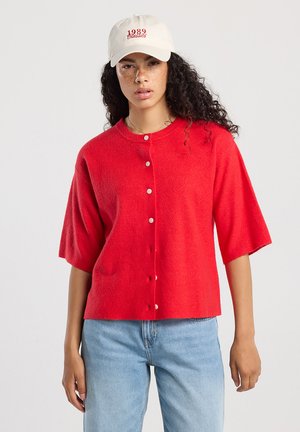 Femme aux cheveux bouclés portant une casquette blanche et un pull à manches courtes rouge à boutons, associé à un jean bleu clair, sur un fond uni.
