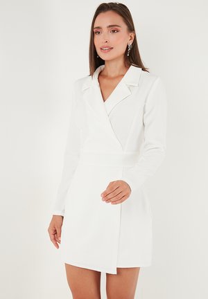 Frau trägt ein weißes Blazer-Kleid mit langen Ärmeln, breitem Kragen und Wickel-Front, stehend vor einem schlichten weißen Hintergrund.