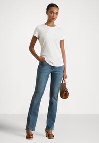 Sieviete baltā izšuvuma t-kreklā, zilās džinsās, brūnās espadrilēs ar platformu, tur rokā brūnu ādas rokassomu, stāvot pret vienkāršu fonu.