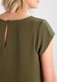 Blouse vert olive avec un tissu texturé, des manches courtes et un dos en trou de serrure avec une petite fermeture à bouton.
