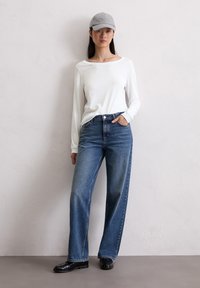Top bianco a maniche lunghe con scollatura a barca, jeans a vita alta azzurri chiari a gamba larga e scarpe nere, complementati da un berretto grigio ricamato.