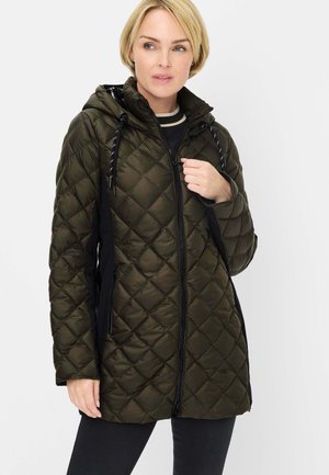 Woman wearing a long, dark green quilted jacket with a hood, black zipper, and black side panels, posing against a plain background.