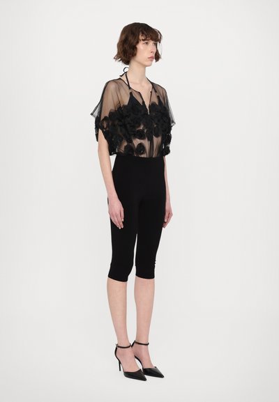 Schwarzes durchsichtige Oberteil mit floralen Akzenten, kombiniert mit schwarzen knielangen Leggings und spitzen Absatzschuhen mit Knöchelriemen.