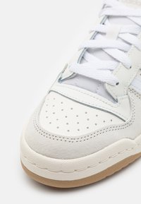 Bílé kožené a semišové tenisky s perforovanou špičkou, plochými bílými tkaničkami, šedými akcenty a textured hnědou gumovou podrážkou.