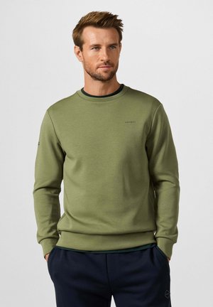 Man met lichtbruin haar, gekleed in een olijfgroene lange mouwen trui en een donkerblauwe broek, staand met handen in de zakken.