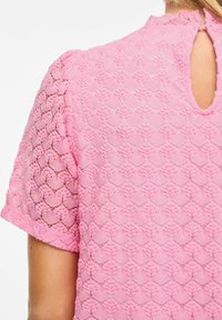 Blouse en dentelle rose avec un motif texturé en losange, manches courtes, et une ouverture en forme de goutte d'eau dans le dos. Légèreté, couture décorative.